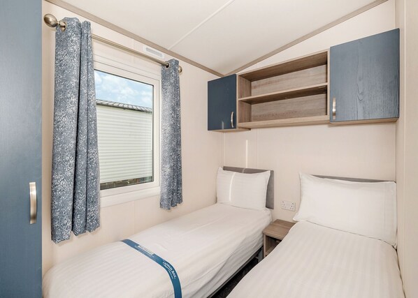Una habitación compacta con dos camas, una ventana con cortinas, un pequeño estante de madera y un enchufe.