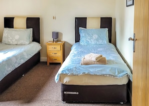 A bedroom with two beds, each with a headboard and pillow, a bedside table with a lamp, and a small drawer. - Lodge with fantastic contemporary styling. (Llanddewi, Nr. Llandrindod Wells)