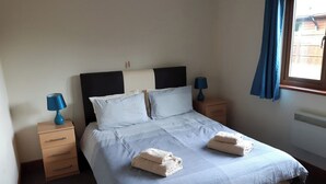 A bedroom with a bed, two bedside tables, two lamps, a window with curtains, and a radiator. - Lodge with fantastic contemporary styling. (Llanddewi, Nr. Llandrindod Wells)