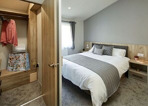 A bedroom with a bed, a nightstand, a wooden wardrobe, and a door leading to another room. - Pondview Retreat Lodge (Glenluce, Newton Stewart)