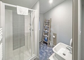 A bathroom with a shower, a sink, a toilet, and a shelf with decorative items. - Pondview Retreat Lodge (Glenluce, Newton Stewart)