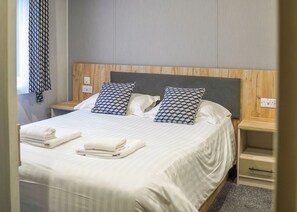 A neatly made bed with white linens and two patterned pillows, a wooden headboard, bedside tables with outlets, and a window with a patterned curtain.