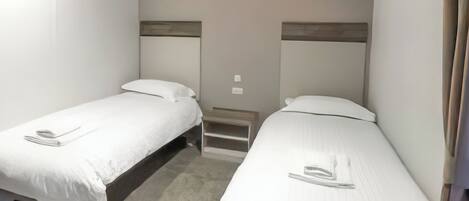 Une chambre double avec deux lits simples, une petite table de chevet et une étagère fixée au mur.