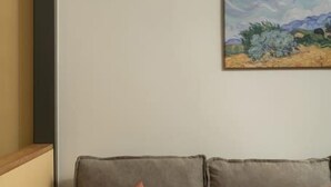 Un salón moderno con un sofá gris, dos cojines naranjas, una lámpara de pared redonda y un cuadro de un paisaje.