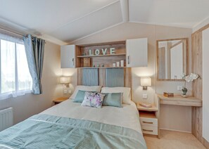 A bedroom with a bed, bedside tables, a nightstand, a mirror, a shelf with decorative items, and a window with curtains.