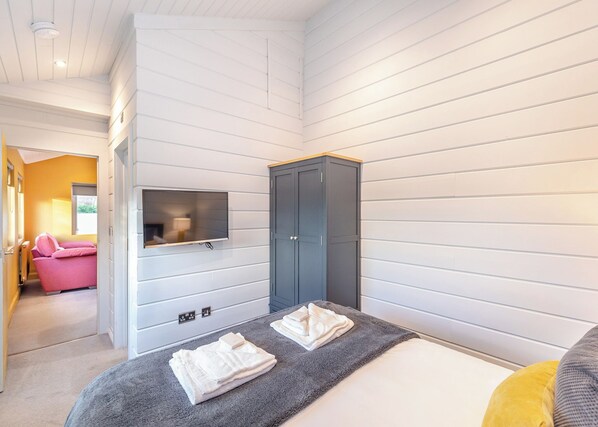 A bedroom with a bed, a grey wardrobe, a TV mounted on the wall, and a pink chair in the adjacent room.