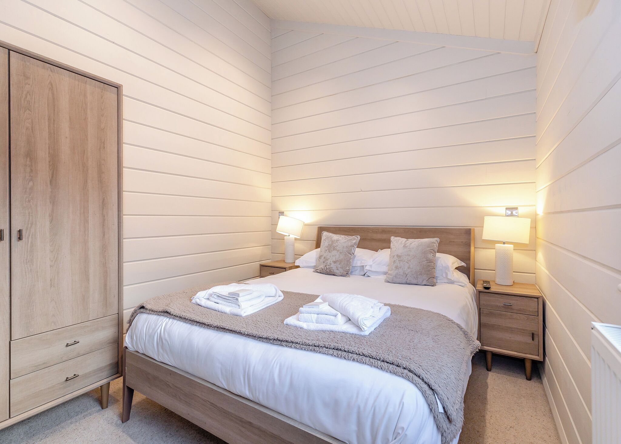 A bedroom with a wooden wardrobe, a bed with white linens, two bedside lamps, and a nightstand with a lamp.
