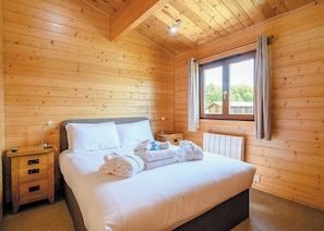 A wooden-paneled bedroom with a bed, nightstands, a window, and a radiator. - Luxurious solid log lodge. (Wenhaston, Halesworth)