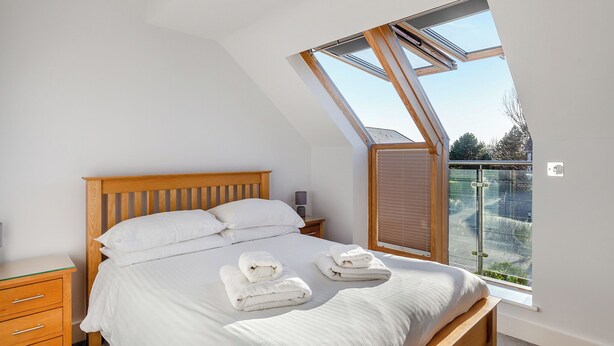 A bedroom with a sloped ceiling, a wooden bed frame, white bedding, and a large window with a view of the outdoors.