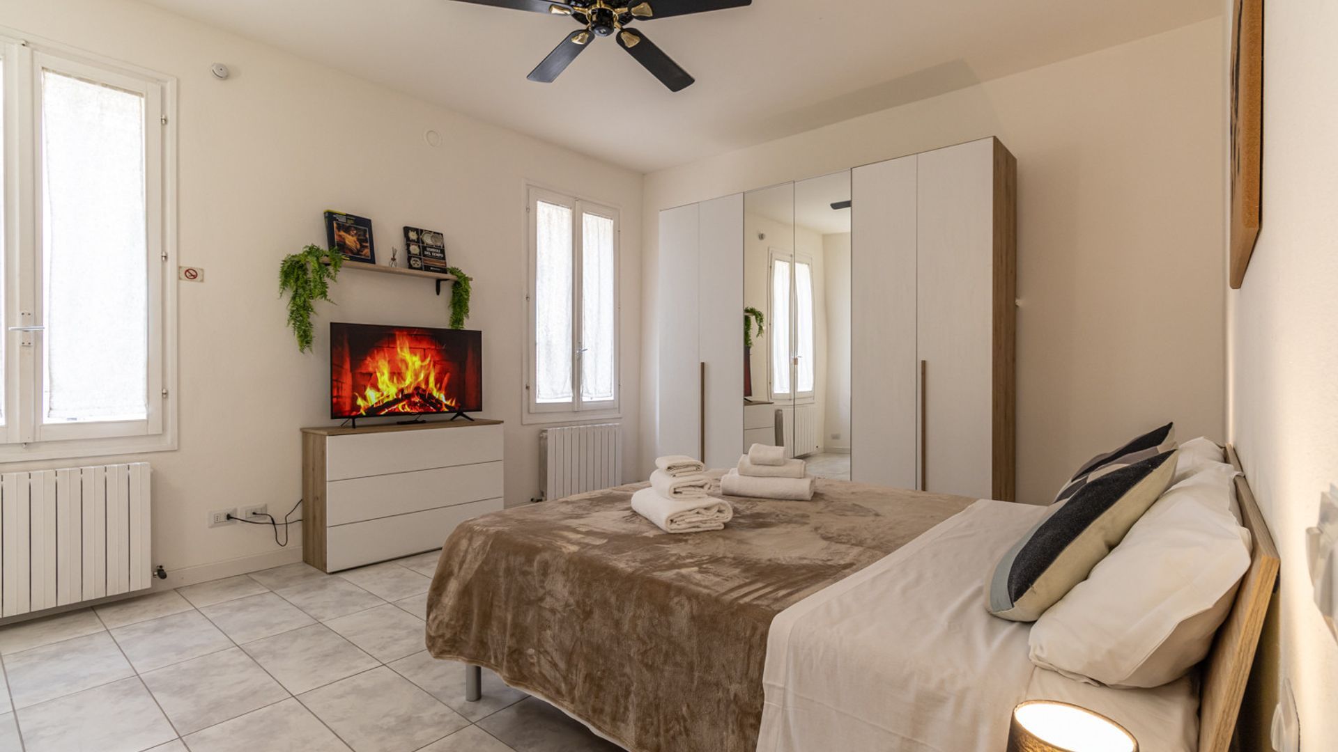 A bedroom with a bed, a TV displaying a fireplace, a wardrobe, and a mirror.