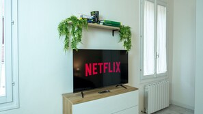 Televisão de tela plana presa na parede, com um console de madeira embaixo, prateleira acima com livros e uma planta, e uma janela à direita.