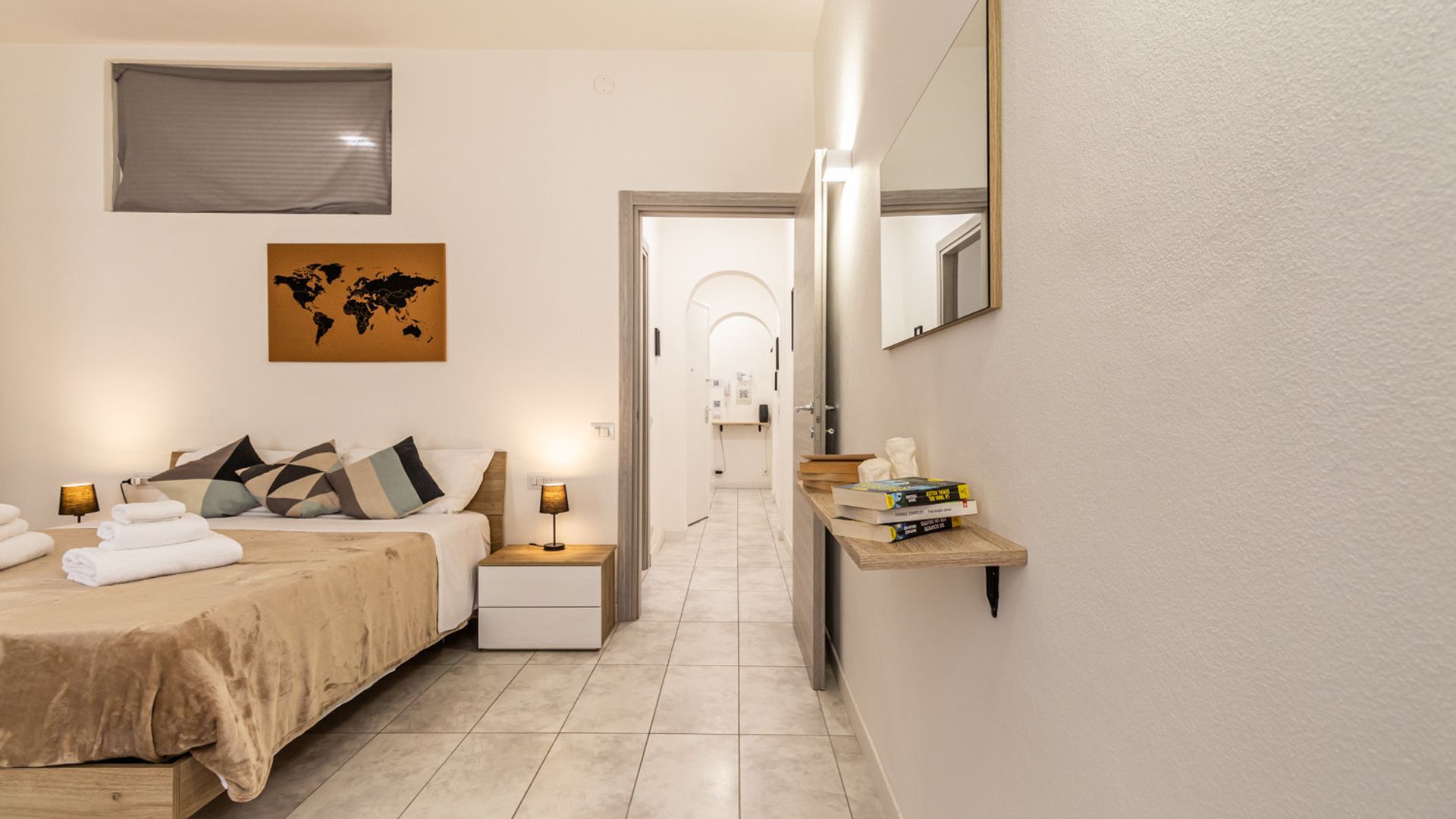 A hotel room with a bed, a bedside table, a lamp, a world map on the wall, a shelf with books, and a mirror.