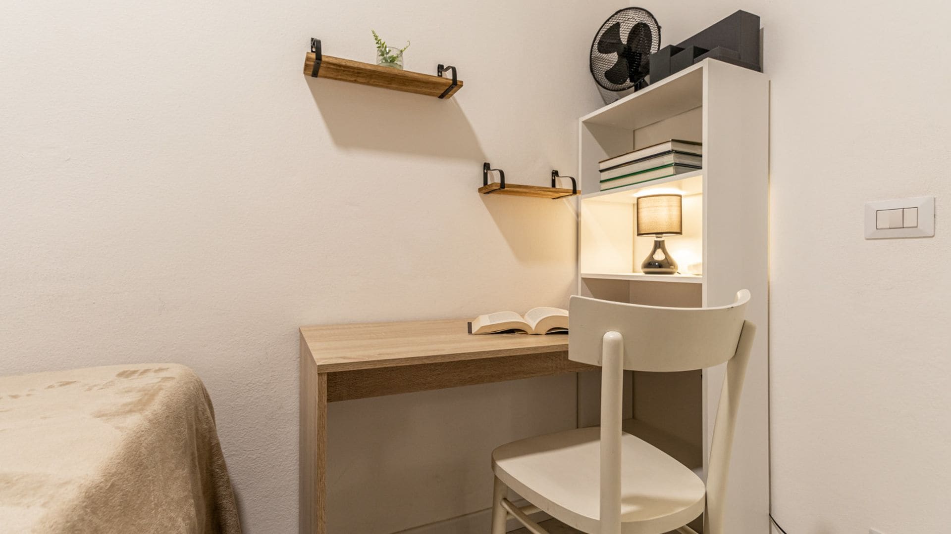 A small, cozy room with a wooden desk, a chair, a shelf with books, a lamp, and a fan.