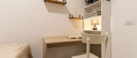 A small, cozy room with a wooden desk, a chair, a shelf with books, a lamp, and a fan.
