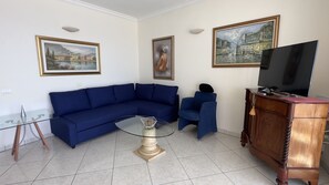 Ein Wohnzimmer mit einem blauen Sofa, einem Glastisch, einer Holzkommode und einem Fernseher.