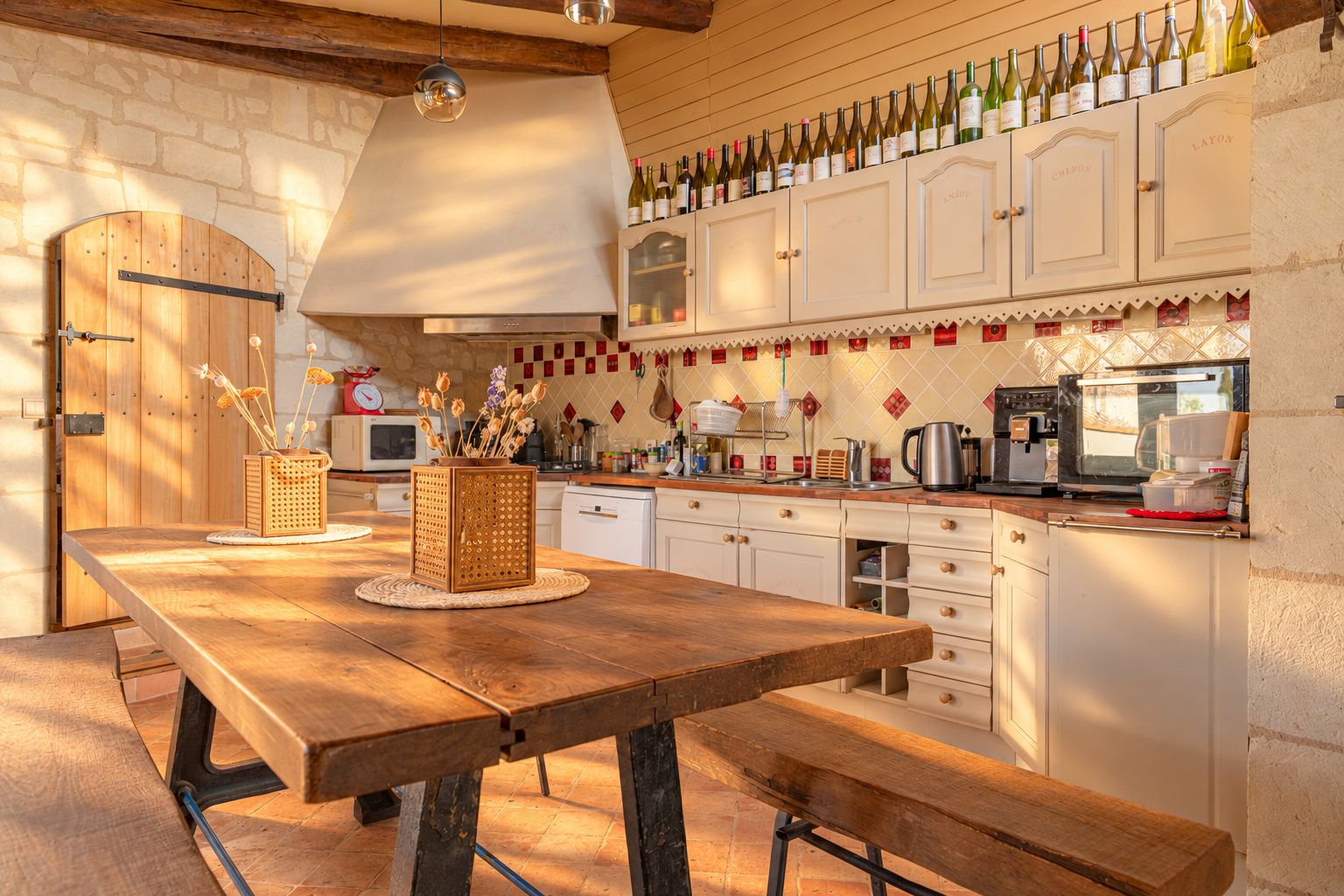 A rustic kitchen with wooden cabinets, a long wooden table, and a wine rack above.
