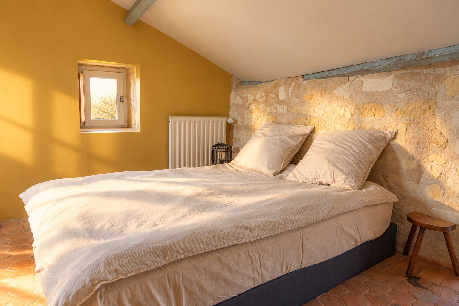 A bedroom with a stone wall, a bed with white bedding, a small window, and a radiator.