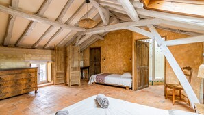 A rustic bedroom with exposed wooden beams, a stone wall, a bed with a rolled towel, a wooden dresser, and a chair.