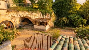 Un bâtiment en pierre avec un toit recouvert de tuiles, une petite structure en bois et un escalier menant à un balcon qui donne sur un jardin verdoyant et une zone pavée.