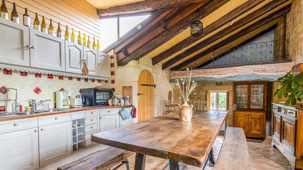 A rustic kitchen with a long wooden table, white cabinets, and a stone wall.
