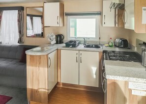 Une petite cuisine équipée comprenant un évier, une cuisinière et des placards, une fenêtre avec des rideaux et un canapé dans l’espace salon.
