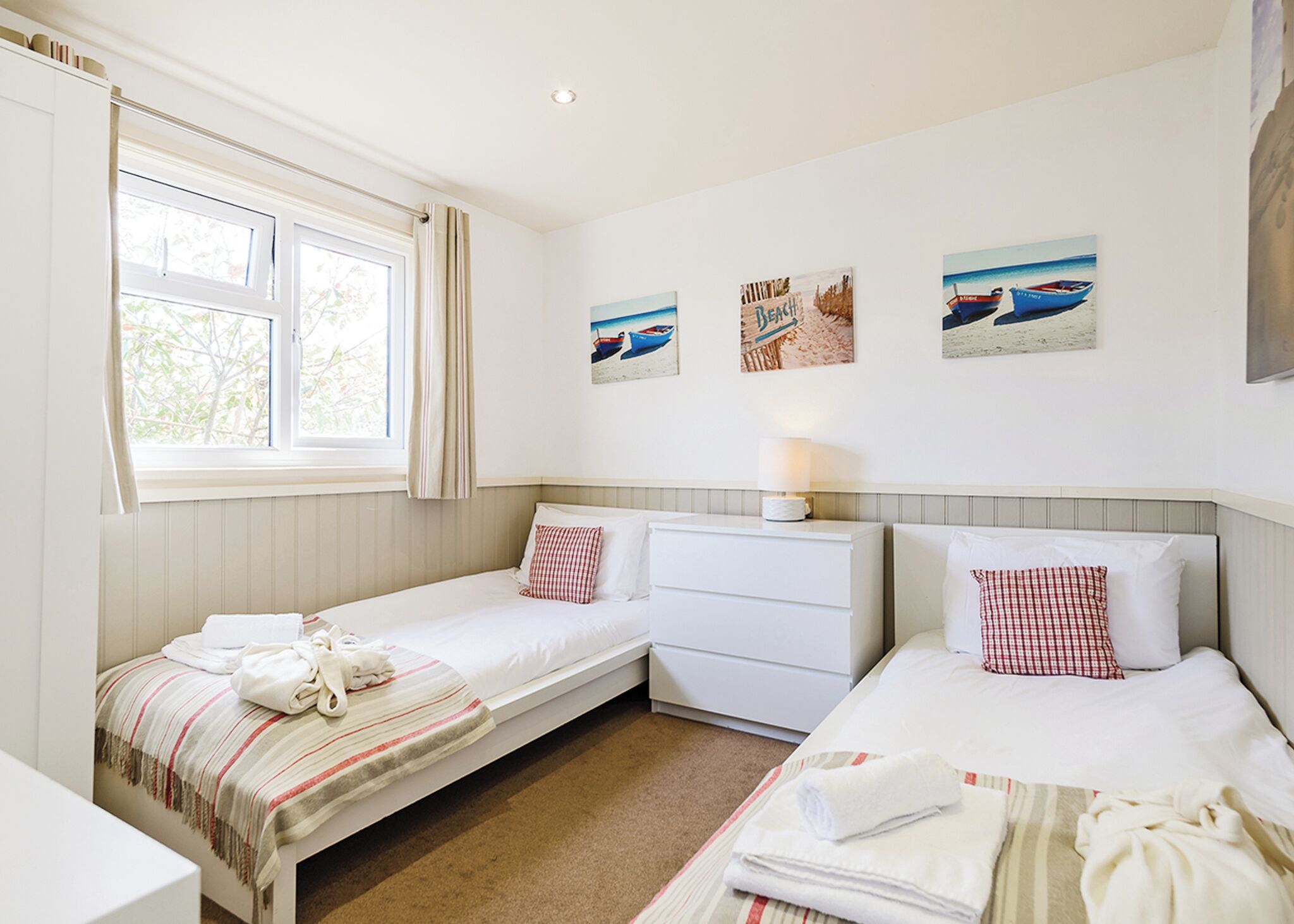 Une chambre avec deux lits simples, une petite commode blanche, une fenêtre avec des rideaux et des œuvres d’art murales représentant des scènes de plage.