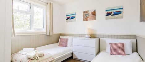 A room with two single beds, a small white dresser, a window with curtains, and wall art depicting beach scenes.