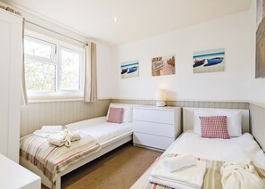 A room with two single beds, a small white dresser, a window with curtains, and wall art depicting beach scenes.