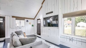 Ein Zimmer mit grauem Sofa, einem kleinen Tisch, einem an der Wand befestigten Fernseher und einem Fenster mit Blick ins Freie.
