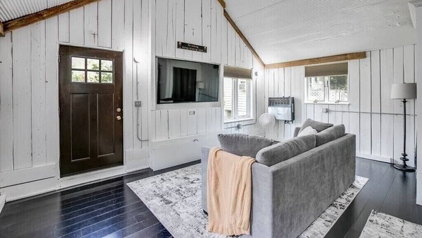 Ein Zimmer mit einer dunklen Holztür, einem grauen Sofa mit Decke und einer weißen Decke mit Holzbalken, sowie einem an der Wand befestigten Fernseher.