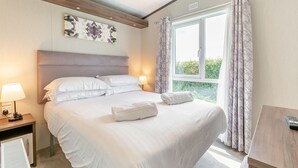 A neatly made bed with white linens, a bedside lamp, a window with curtains, and a wall-mounted artwork.