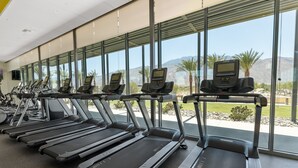 Varias caminadoras con pantallas digitales en un gimnasio con amplios ventanales que dan a un paisaje desértico.