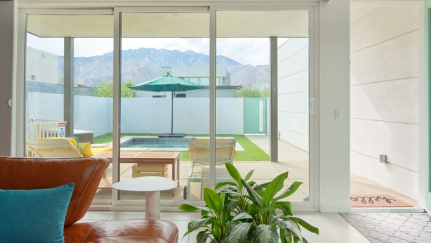 Una sala de estar moderna con un suelo de vidrio corredera que da a un patio con piscina, mobiliario exterior y vistas a la montaña.
