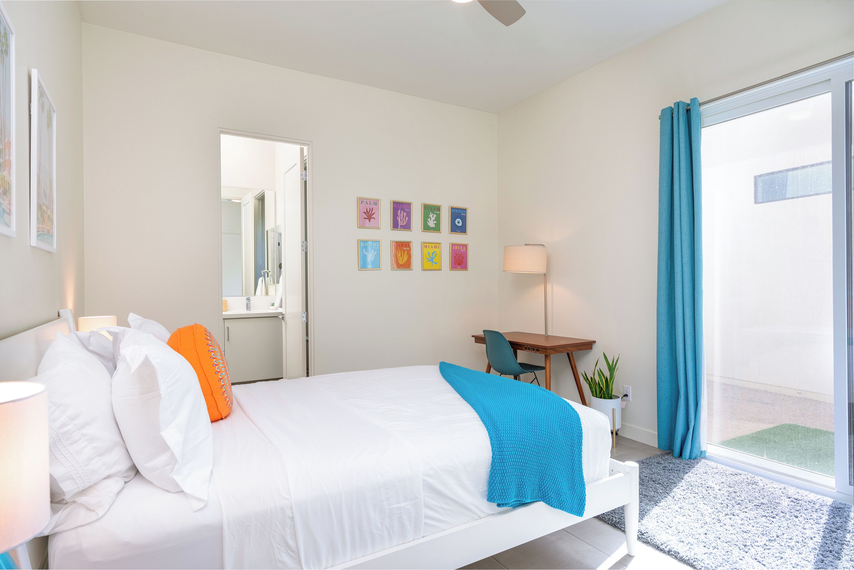 Habitación de hotel con cama, escritorio, silla, lámpara, ventana con cortinas y pared con cuadros.