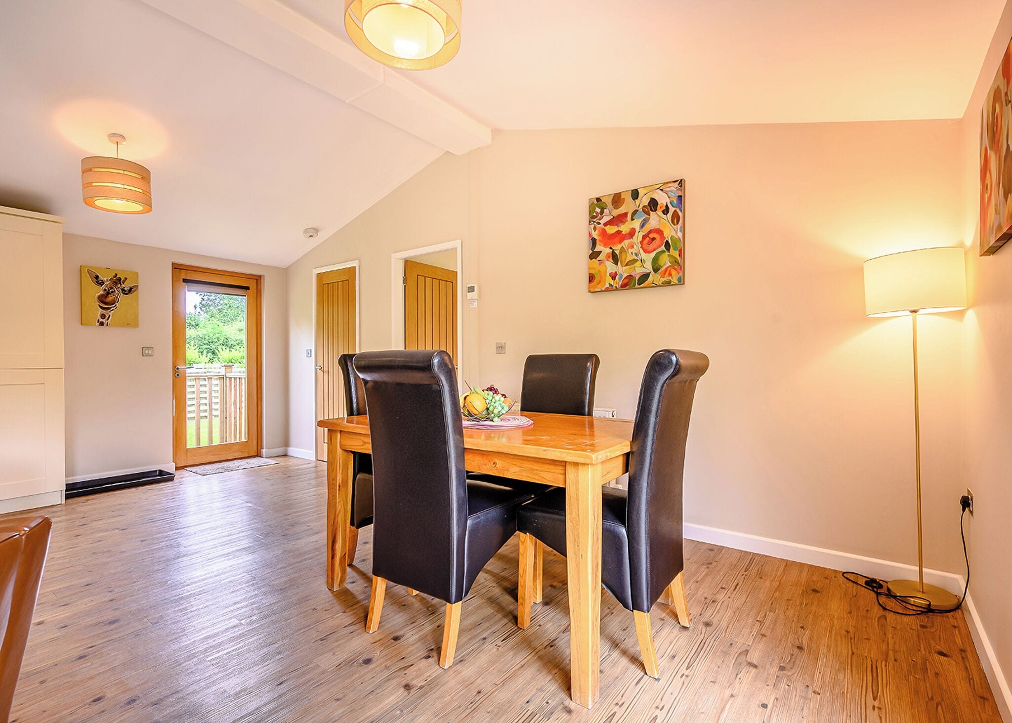 A dining area with a wooden table and four chairs, a painting on the wall, and a door leading to a balcony.