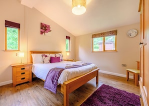 A bedroom with a wooden bed, two bedside tables, a nightstand, a purple rug, and a window with a view of greenery.