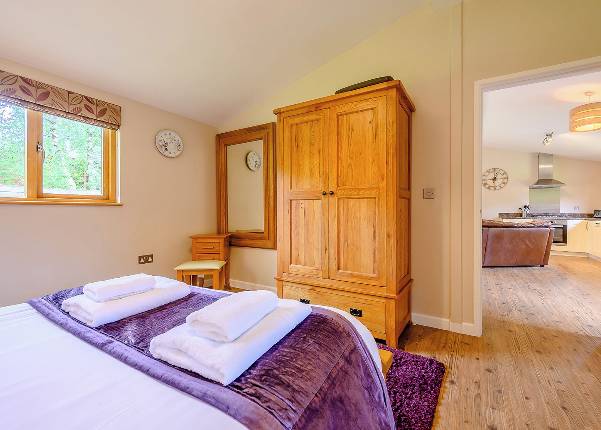 A bedroom with a bed, a wooden wardrobe, a mirror, a bedside table, and a window.