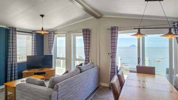 A living room with a sofa, TV, dining table, and chairs, large windows with curtains, and a view of the sea.