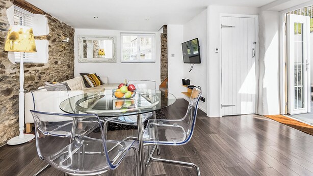A modern living room with a glass dining table, clear chairs, a stone accent wall, a sofa, and a TV mounted on the wall. - Apple Cottage is a firm favourite with our guests. (Watergate, Looe)