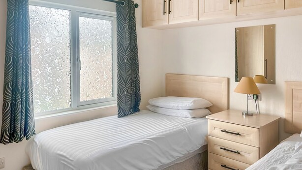A bedroom with a bed, a nightstand, a lamp, a dresser, and a window with curtains. - Country Lodge Four Spa (Tintagel)