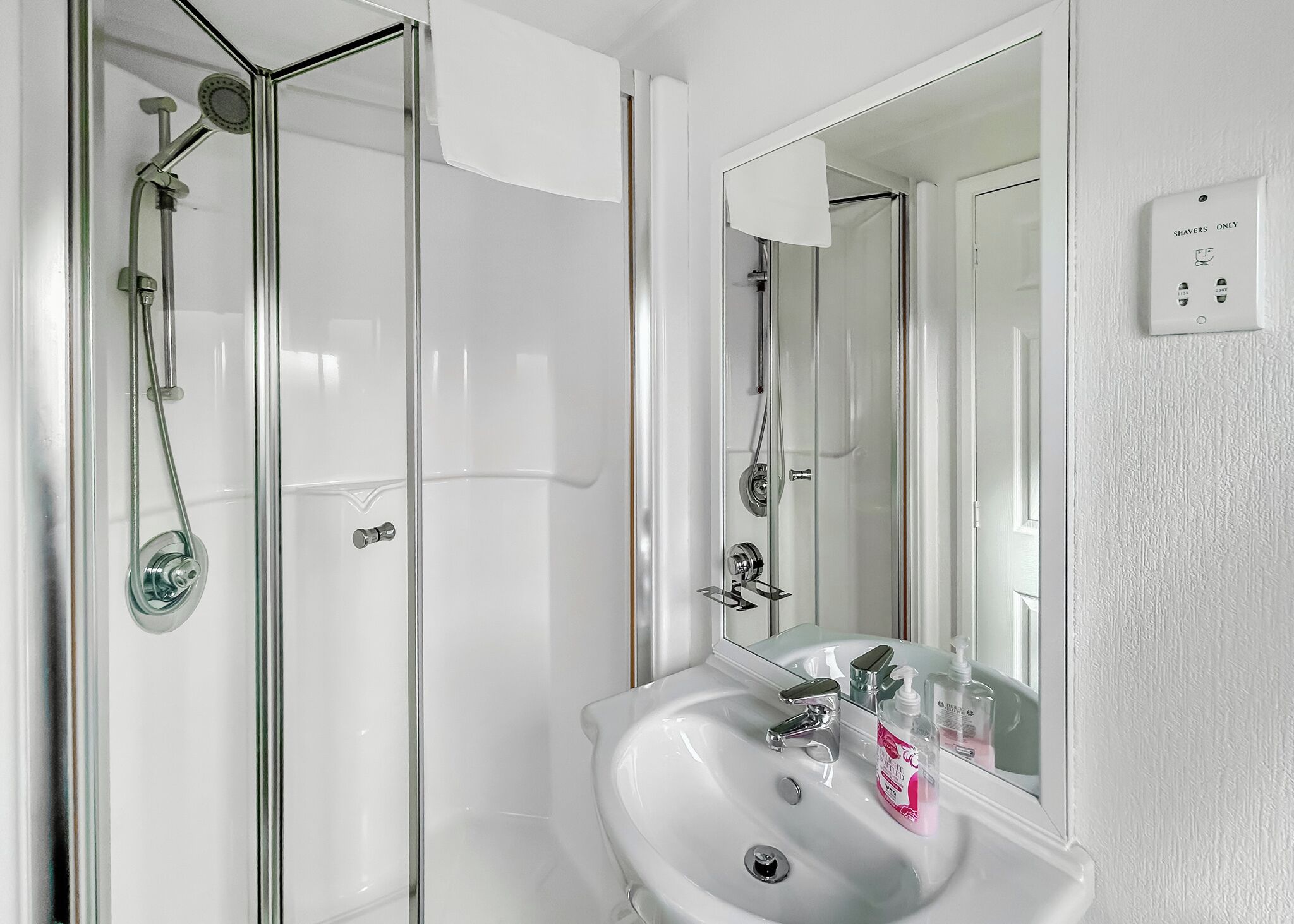 A bathroom with a glass shower enclosure, a white sink with a mirror, and a 'Shavers Only' sign.