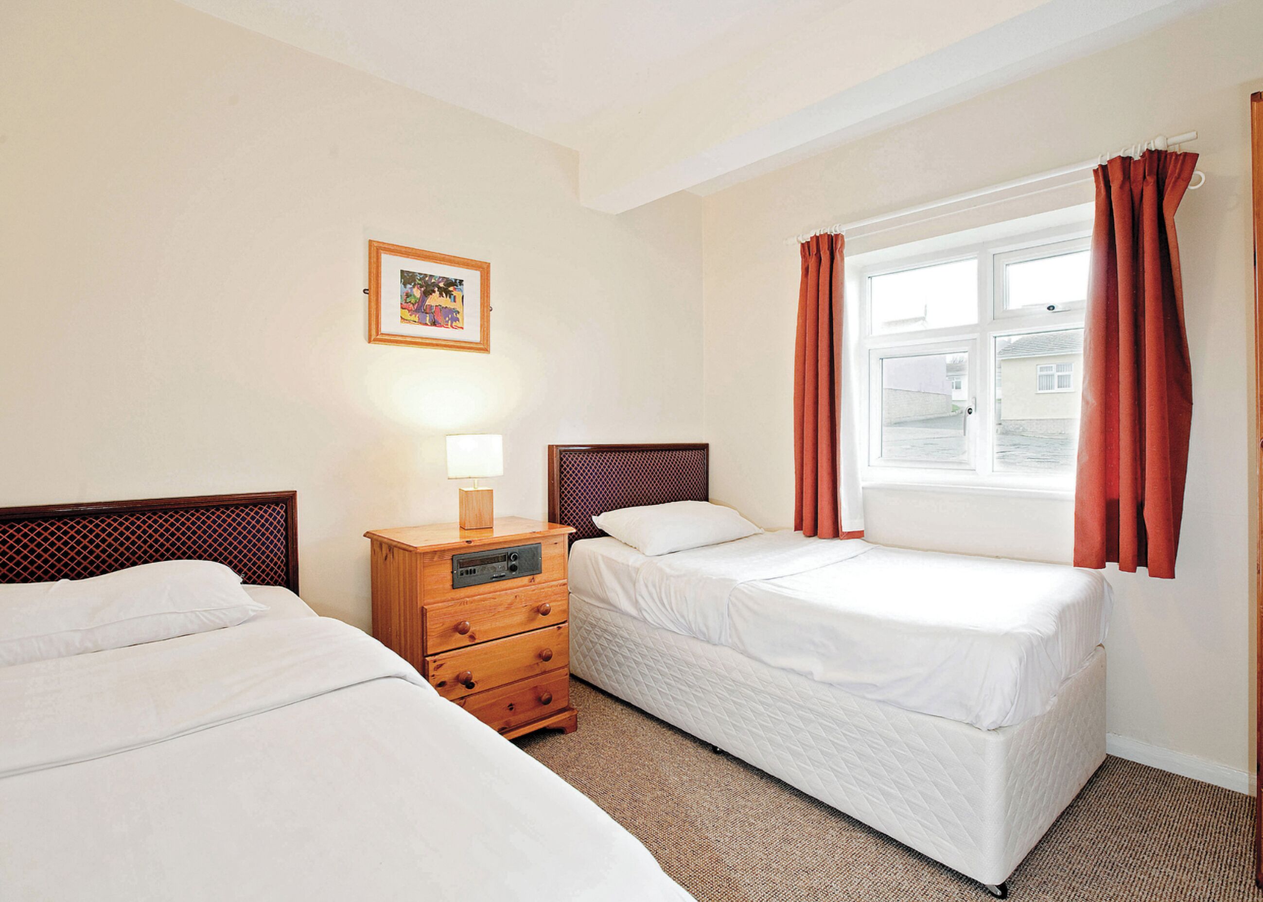 A hotel room with two beds, a wooden nightstand with a lamp, a framed picture on the wall, and a window with curtains.