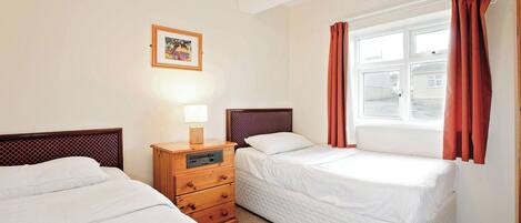 Habitación de hotel con dos camas, una mesita de noche de madera con una lámpara, un cuadro enmarcado en la pared y una ventana con cortinas.