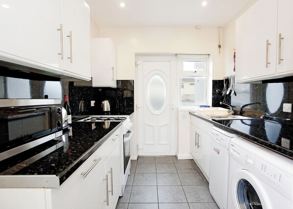 A modern kitchen with white cabinetry, a black countertop, a built-in oven, and a washing machine. - Bungalow with one double with wash basin, one double and one twin. (Trearddur Bay, Holyhead)