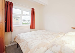 Quarto com cama, janela com cortinas, radiador e vista para uma casa.