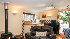 A cozy living room with a fireplace, a leather sofa with a 'home' pillow, a kitchen area with a stove, and a view of the outdoors. - The Retreat (Oswestry)