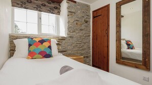 Une chambre avec un mur en pierre, une porte en bois, un lit avec une literie blanche, un coussin coloré et un miroir accroché au mur.