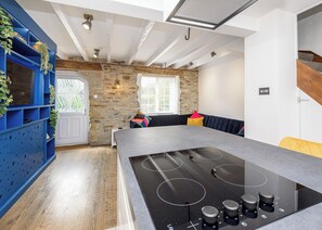 Cozinha moderna com ilha central, prateleiras azuis embutidas, parede de pedra e um espaço de estar acolhedor com almofadas.