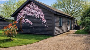 A wooden cabin with a sloped roof, a gravel driveway, and a garden with a small tree and shrub.