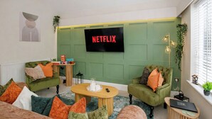 A living room with green walls, a Netflix sign, two green armchairs, a wooden coffee table, and a small side table with a laptop.
